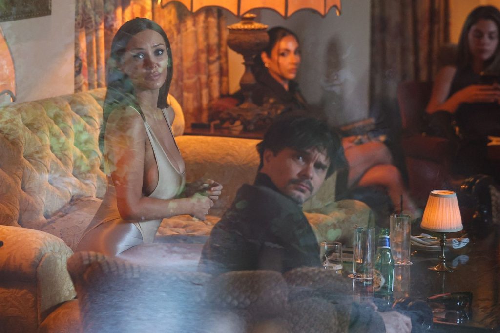 A woman in a revealing dress and a man in dark clothes sit on a vintage sofa in a dimly lit, warmly decorated living room with several other people in the background and drinks on the table.