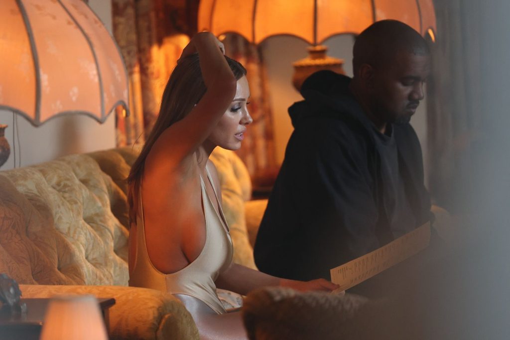 A woman in a sleeveless beige bodysuit sits on a yellow sofa, lifting her arm, while a man in dark clothing sits beside her, both illuminated by warm light from vintage lamps.