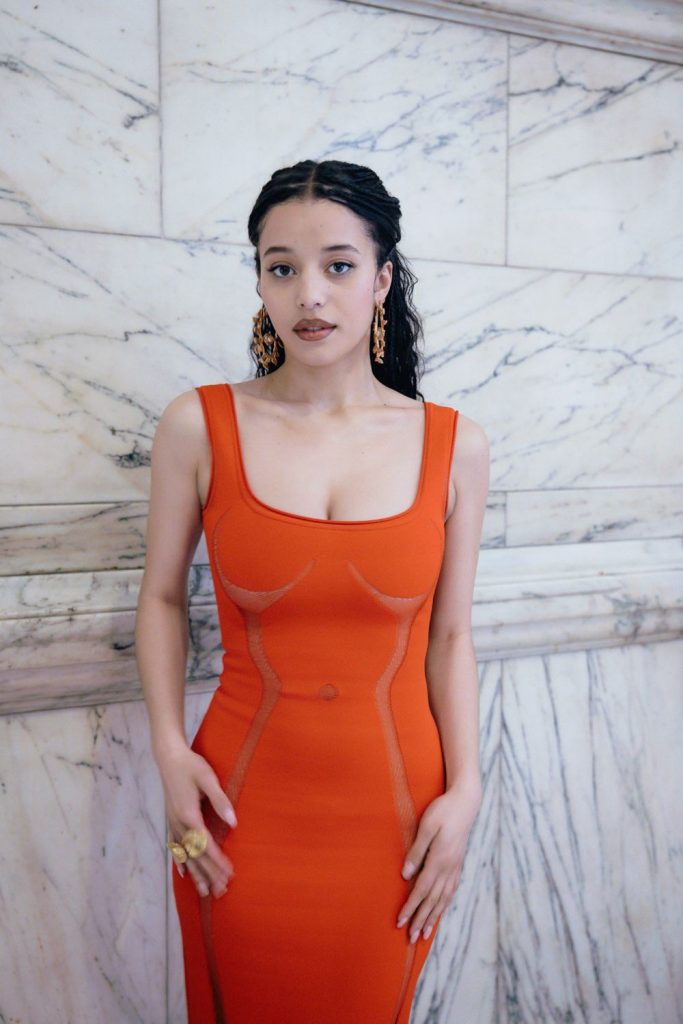 A woman with wavy dark hair and statement earrings wears a form-fitting, sleeveless orange dress, standing in front of a white marble wall with grey veining.