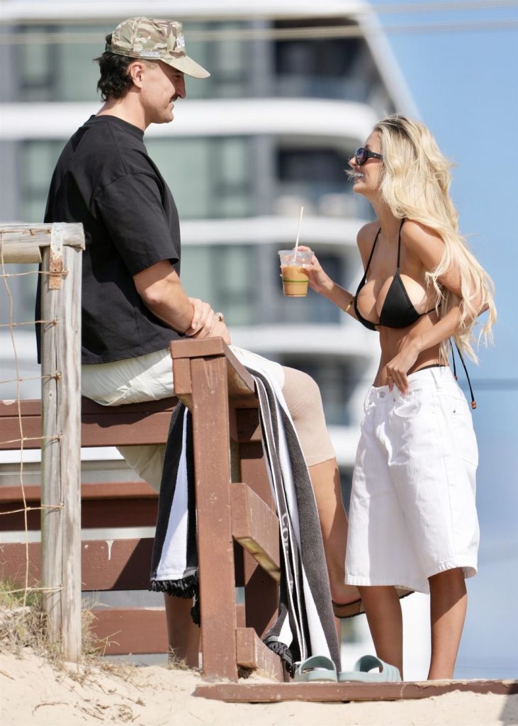 A man sits on a wooden structure at the beach, wearing a cap, black shirt, and shorts. A woman in a black bikini top and white skirt stands beside him, smiling and holding an iced coffee. Buildings are visible in the background.