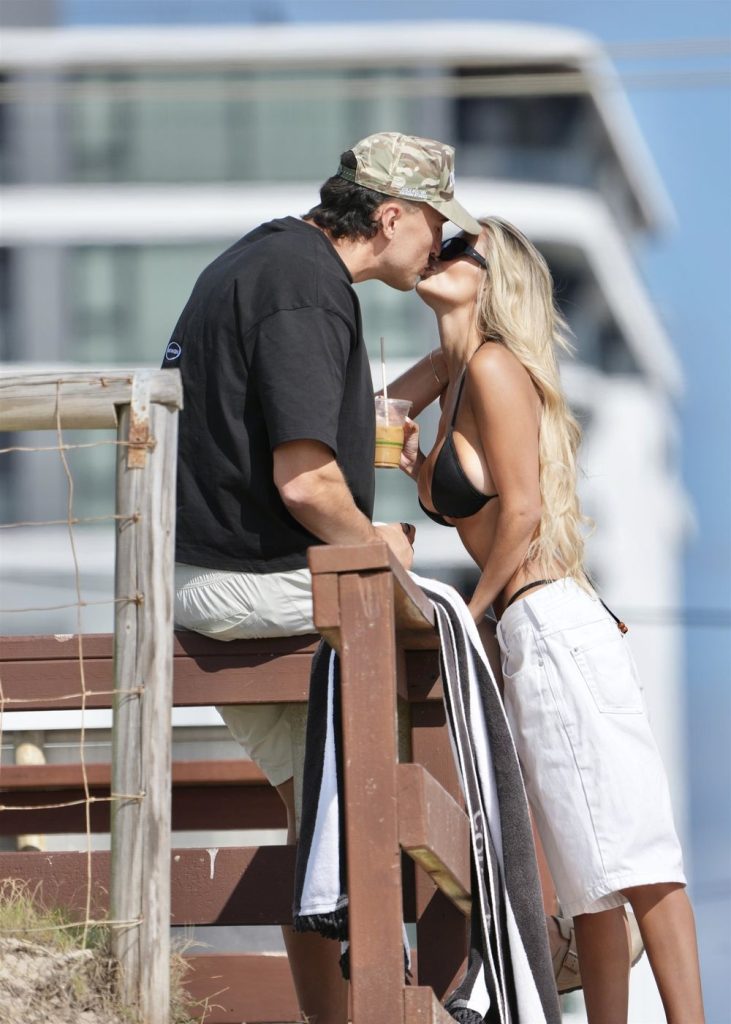 A man in a cap and a woman in a black bikini top and white shorts share a kiss outdoors near a wooden railing, with a modern building blurred in the background. The woman holds a drink in her hand.