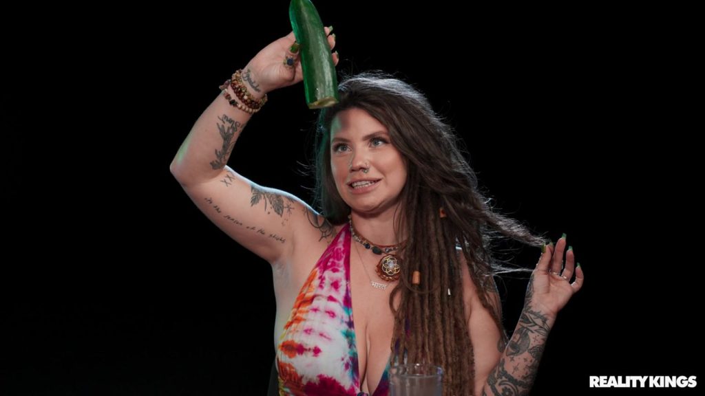 A tattooed woman with dreadlocks in a tie-dye top holds up a cucumber against a black background. She wears layered necklaces and beaded bracelets.