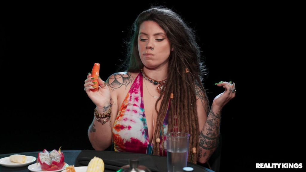 A woman with long dreadlocks and colorful tattoos sits at a table holding a small carrot in one hand. She wears a tie-dye halter top, and there are plates of food and a glass of water in front of her. The background is black.