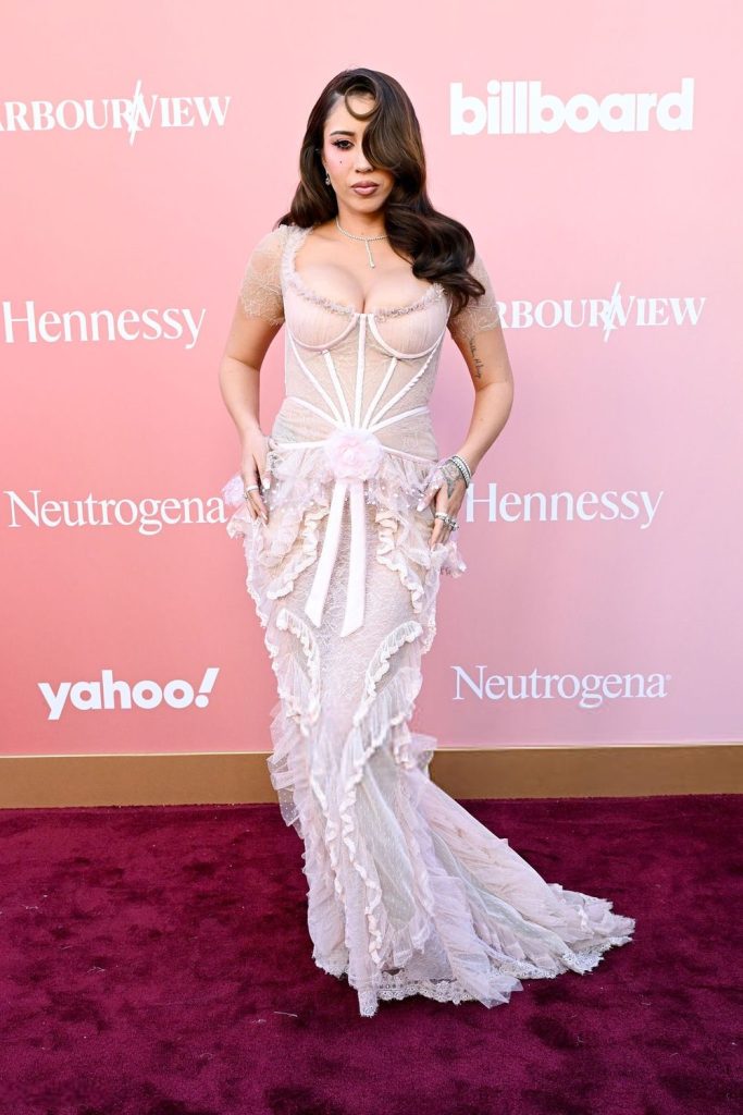 A woman in a pale pink, lace, ruffled gown poses on a red carpet in front of a backdrop with logos including Billboard, Neutrogena, and Hennessy. She has long wavy hair and is wearing statement jewelry.
