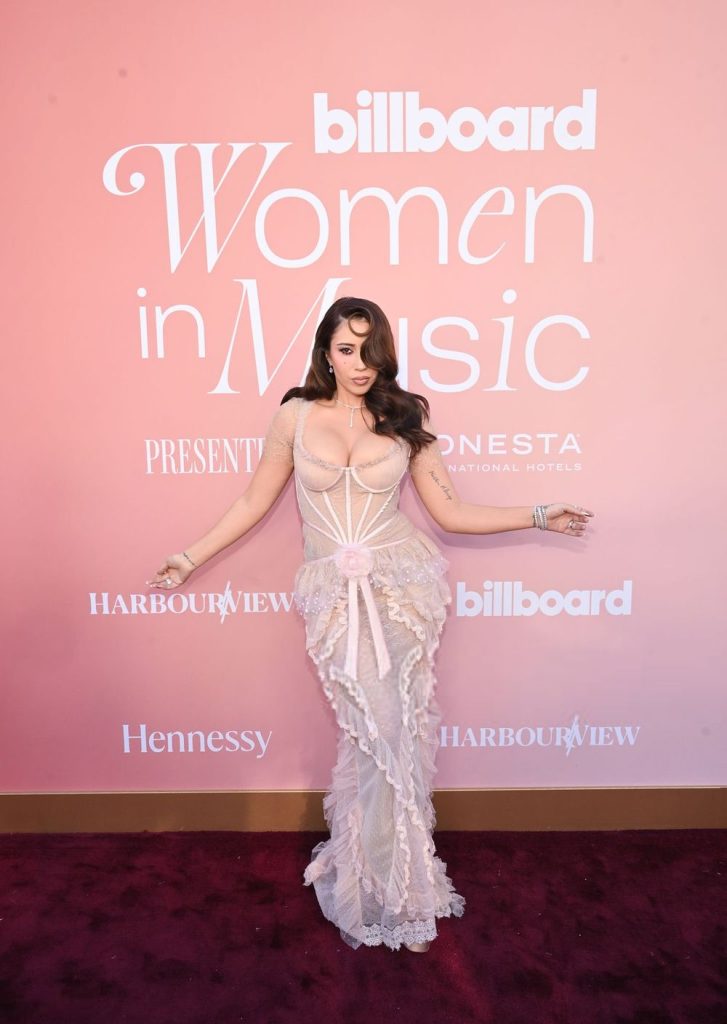 A woman in a sheer, form-fitting, light pink gown poses on the red carpet in front of a pink backdrop with “billboard Women in Music” and sponsor logos.