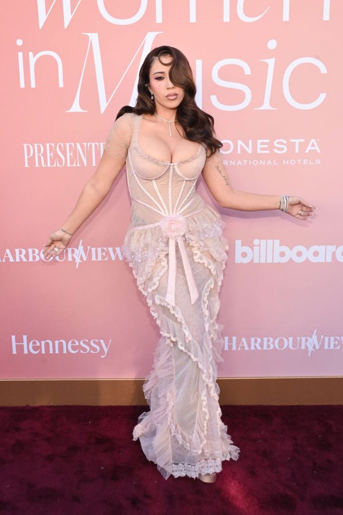 A woman in a sheer, form-fitting, light pink gown poses on the red carpet in front of a pink backdrop with “billboard Women in Music” and sponsor logos #2.