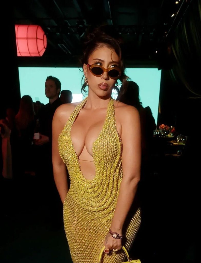 A woman wearing a sparkling, gold halter dress and sunglasses stands confidently at an indoor event, surrounded by people in semi-dark lighting. She holds a matching gold purse and has her hair styled up.