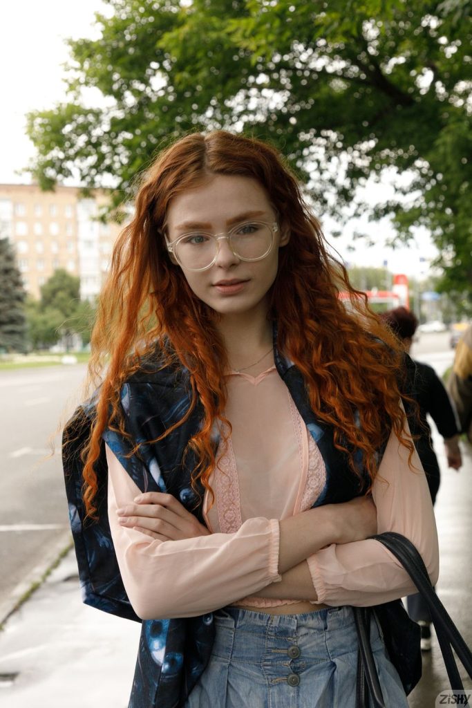 A young woman with long curly red hair and glasses stands outdoors with her arms crossed, wearing a sheer pale pink blouse and light blue high-waisted jeans. Trees and a street are visible in the background.