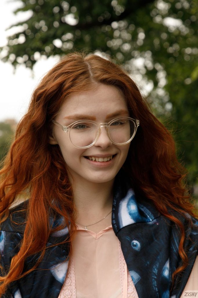 A young woman with long red hair and large round glasses is smiling outdoors. They are wearing a pink top and a blue patterned jacket, with green trees blurred in the background.