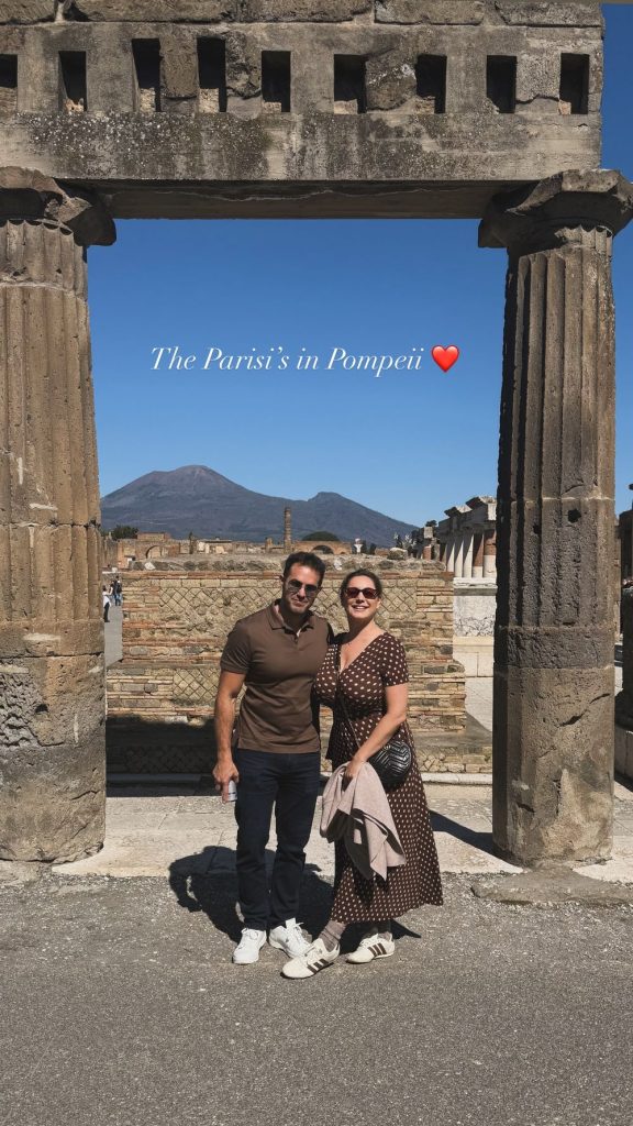 A man and woman stand smiling under a stone archway in Pompeii, with ancient ruins around them and a volcano in the background. The text above reads The Parisis in Pompeii with a red heart emoji.