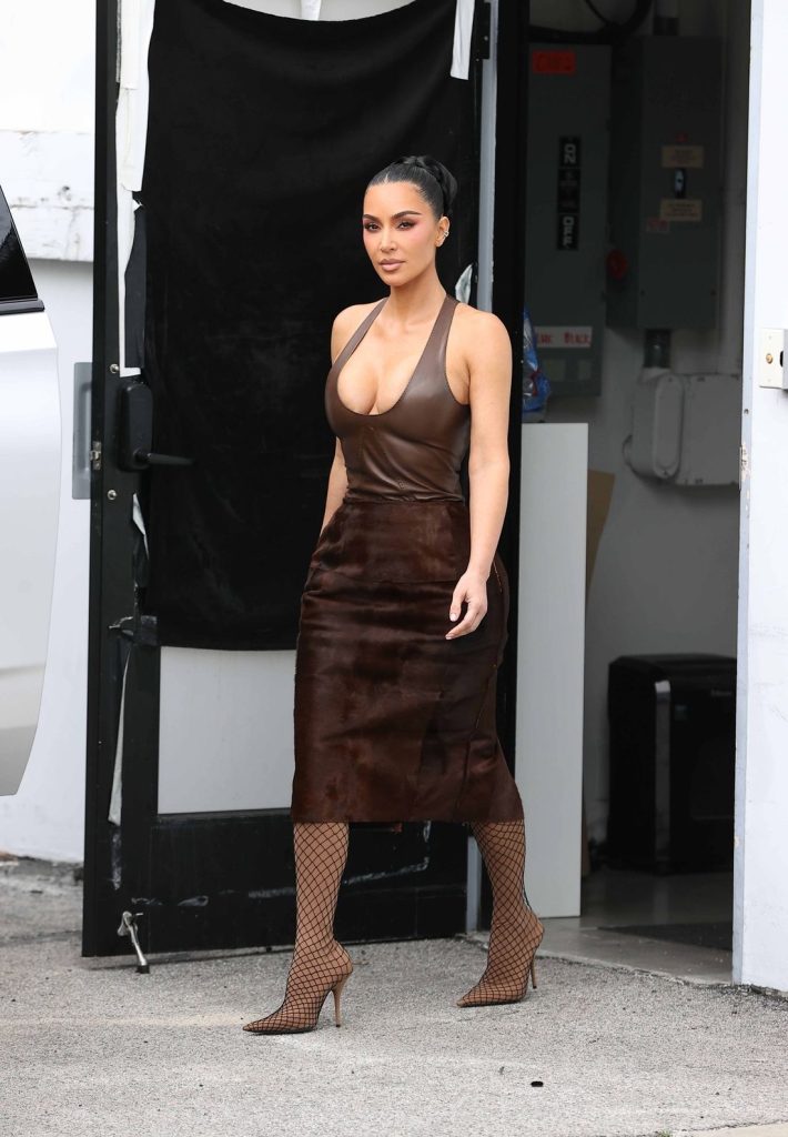 Kim Kardashian with dark hair in a sleek ponytail walks outside wearing a brown halter-style leather dress and fishnet heels. She is confident and stylish, stepping out of a doorway.