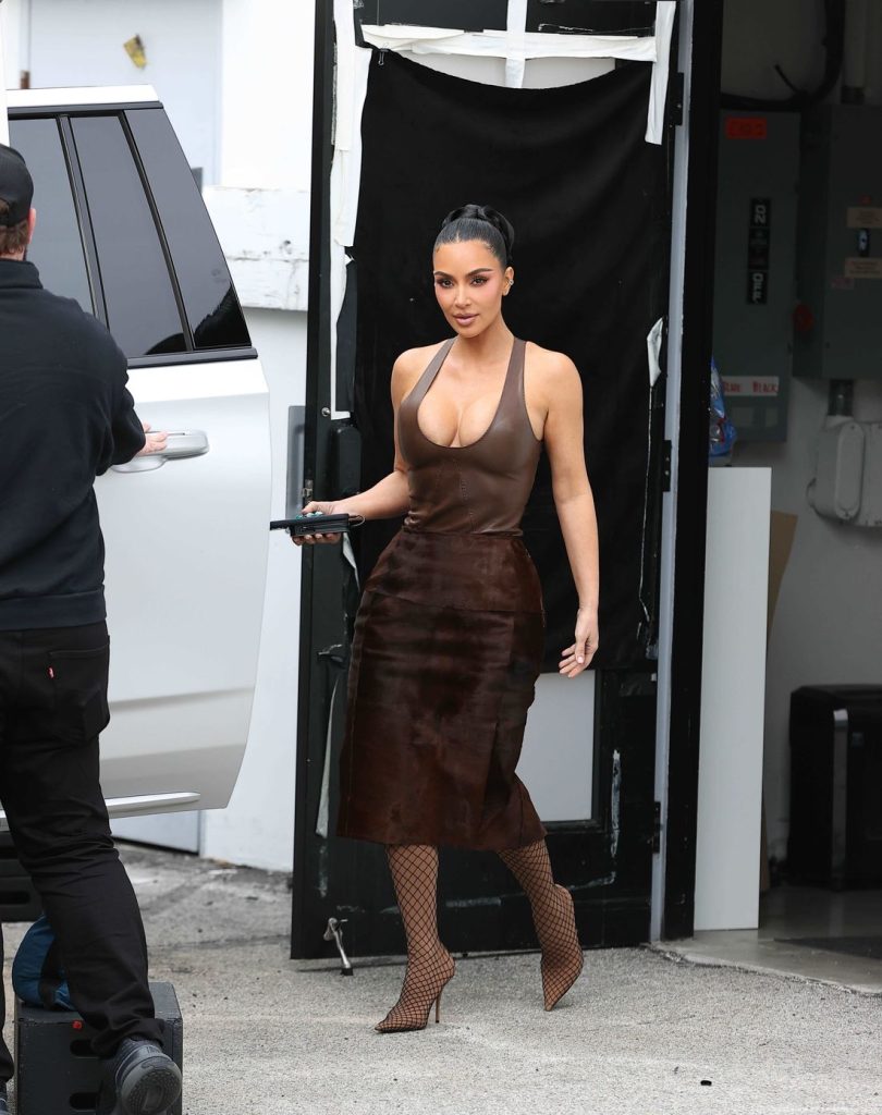 Kim Kardashian is stepping outside through a black doorway, holding a phone and looking forward.