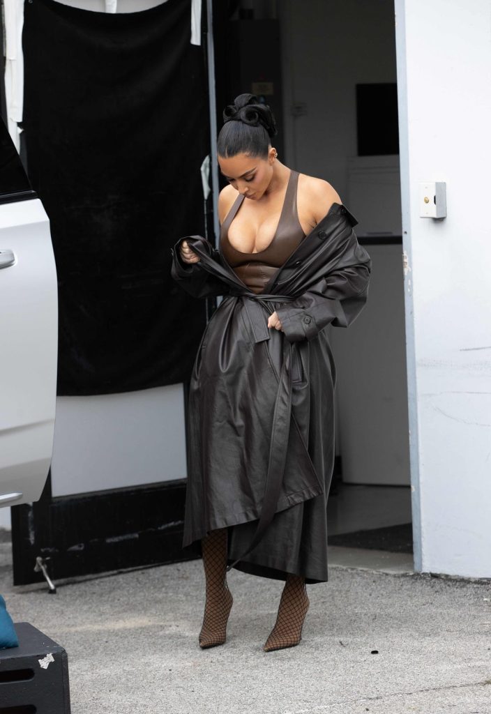 Kim Kardashian stands outside near a white vehicle, looking down while adjusting her coat.