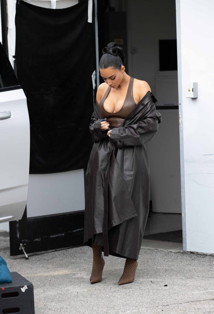 Kim Kardashian stands outside near a white vehicle, wearing a long dark coat over a brown top and matching brown heeled boots, looking down as she adjusts her coat.