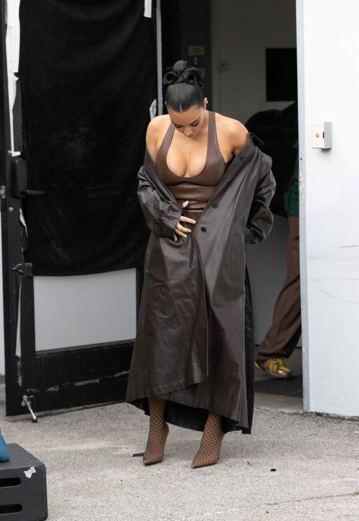 Kim Kardashian wears a long brown leather coat, a brown halter top, a matching skirt, and patterned high heels while standing outdoors and looking down, adjusting her outfit.