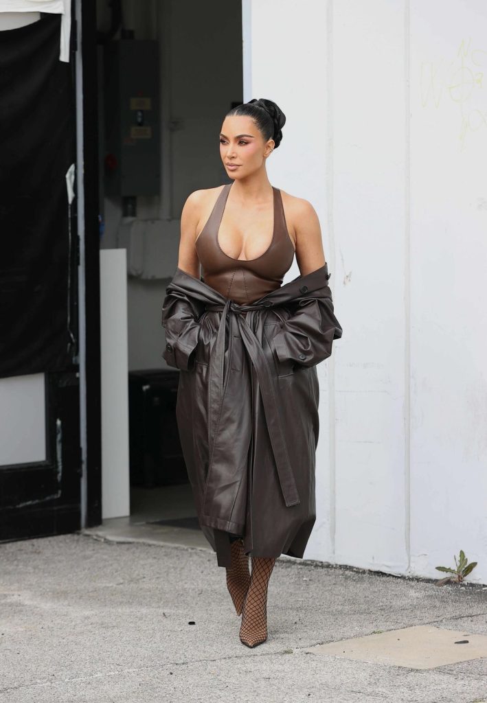 Kim Kardashian with dark hair in a sleek bun wears a brown halter top, a long brown leather trench coat belted at the waist, fishnet boots, and walks outside near a white wall and a black doorway.
