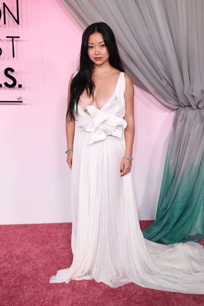 Asian woman with long black hair wearing a white, flowing gown with a large bow detail stands on a pink carpet in front of draped gray curtains and a sign with black text.