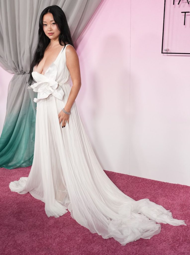 Side view of Asian woman wearing a white, deep plunge gown on a pink carpet in front of a pink background and drapes. She looks straight ahead.