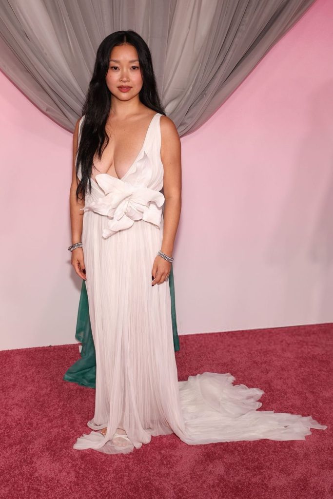 Asian woman with long black hair wears a flowing, white, deep V-neck gown with a large bow at the waist, standing on a pink carpet in front of pink and gray draped fabric.