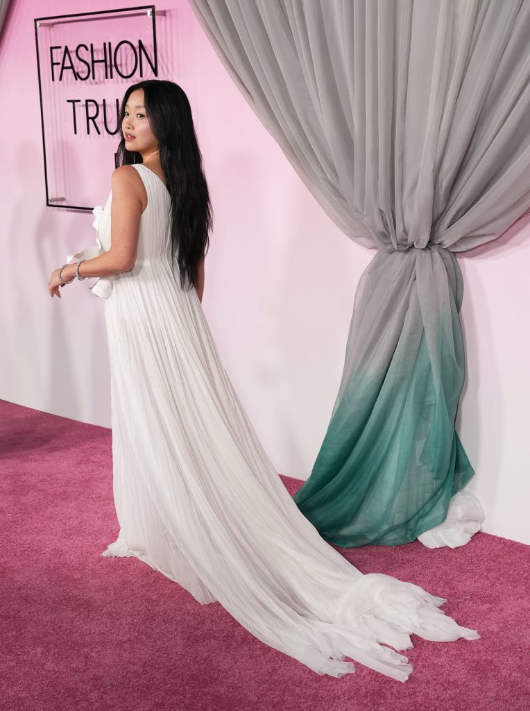 Asian woman in a white gown walking along a pink carpet in front of a pink background and drapes.