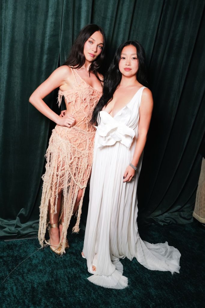 Two women stand in front of a dark green curtain. One wears a peach, fringed dress with sheer details; the other wears a white gown with a large bow at the waist. Both have long, dark hair and neutral expressions.
