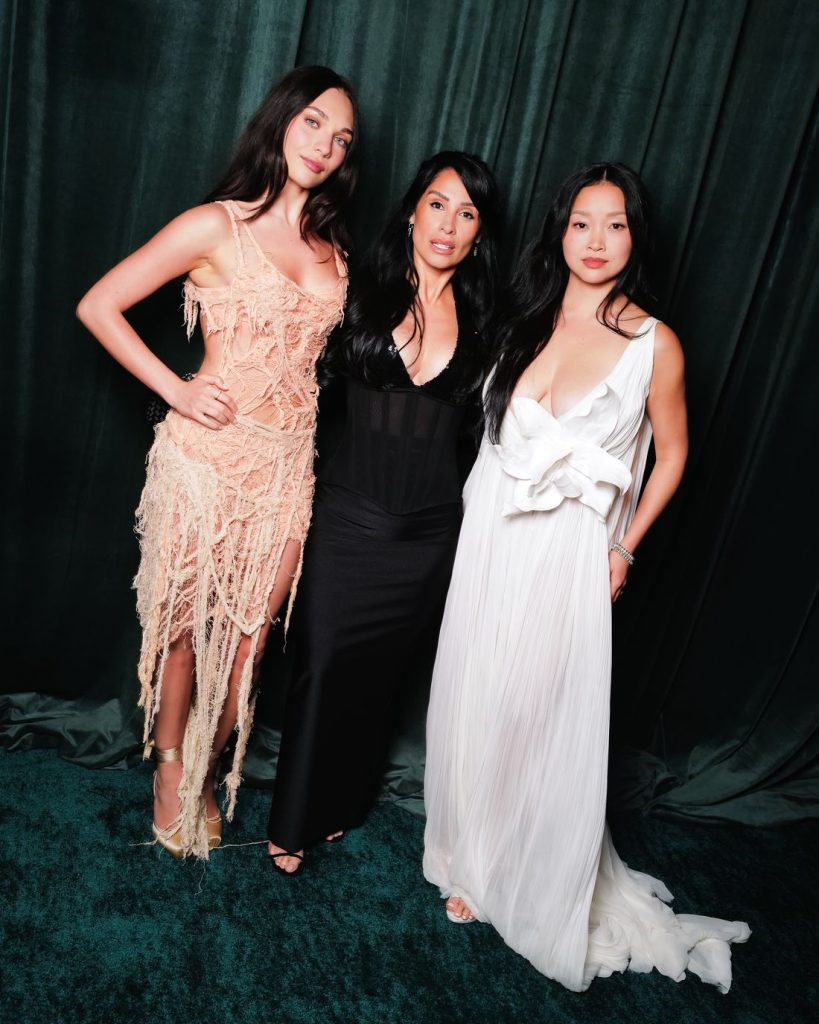 Three women pose together in formal evening gowns against a dark green curtain. The woman on the left wears a peach fringed dress, the center woman is in a fitted black dress, and the woman on the right wears a flowing white gown.