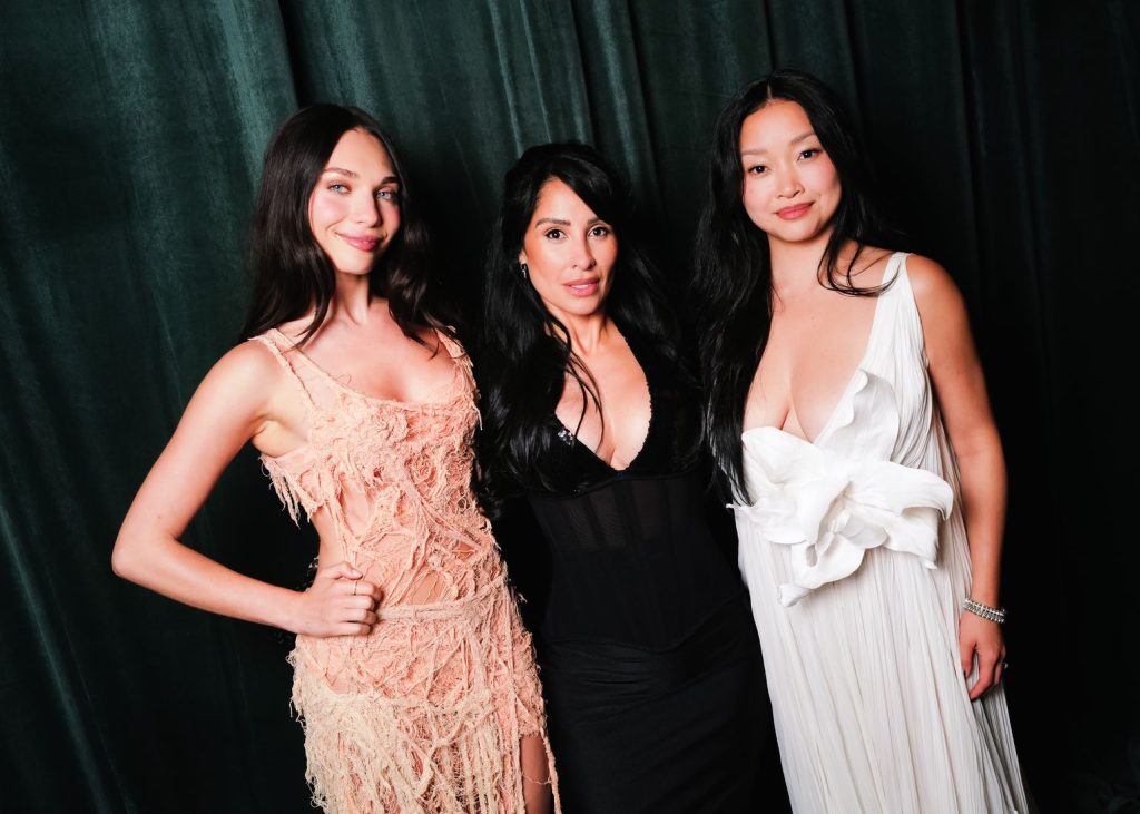 Three women pose together in front of a dark green curtain. One wears a peach lace dress, the middle wears a black dress, and the third wears a white dress with a large bow detail on the front.