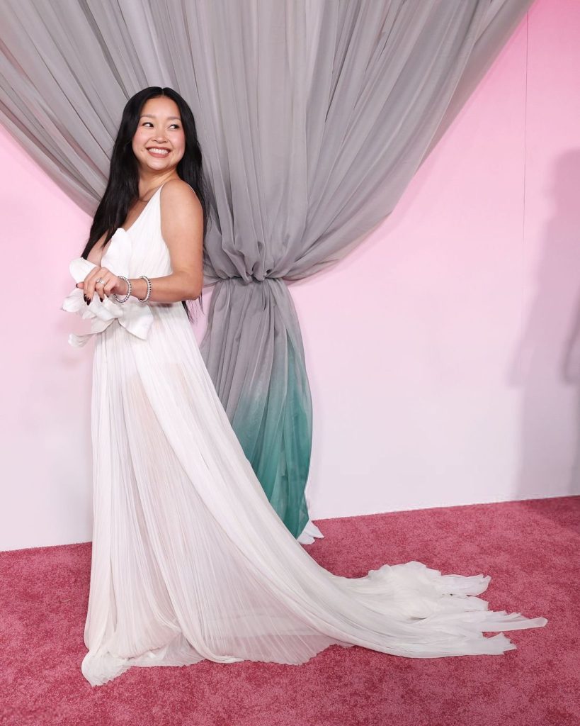 Asian woman in a flowing white gown poses on a pink carpet in front of gray draped curtains, holding the dress’s train and smiling while looking to the side.