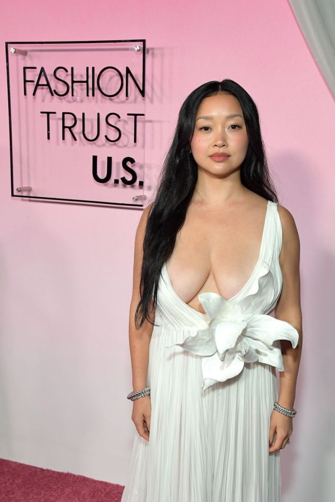 Close up of Asian woman with a slight smile, wearing a low-cut gown in front of a pink background with a sign.