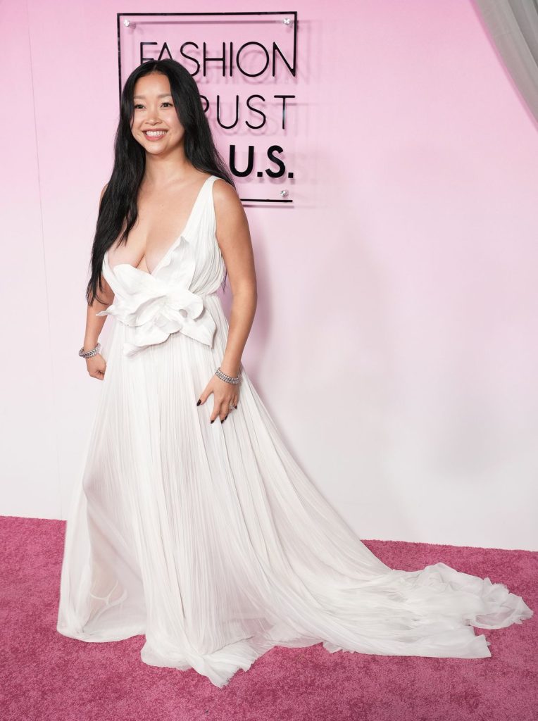 Side view of Asian woman wearing a white, deep plunge gown on a pink carpet in front of a pink background.