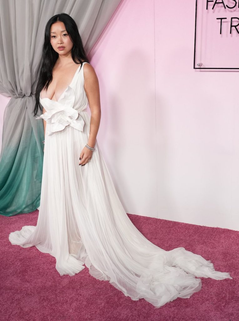 Side view of Asian woman wearing a white, deep plunge gown on a pink carpet in front of a pink background and drapes. She turns her head to face the camera.