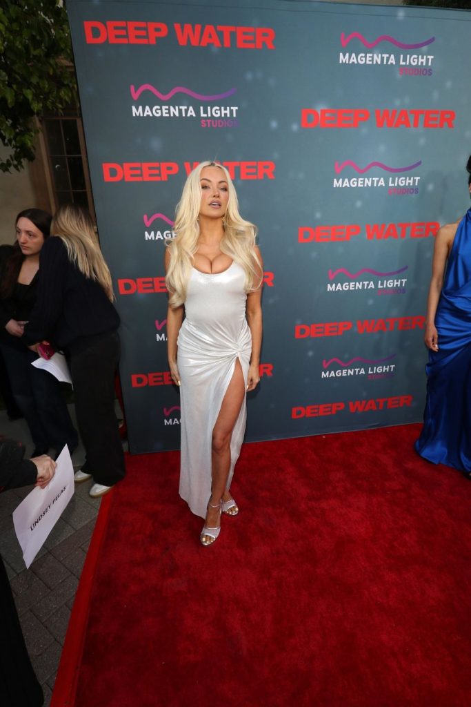 A woman in a white dress with a high slit poses on a red carpet at an event for Deep Water. The backdrop displays logos for Deep Water and Magenta Light Studios in pink and red. Other people stand nearby.