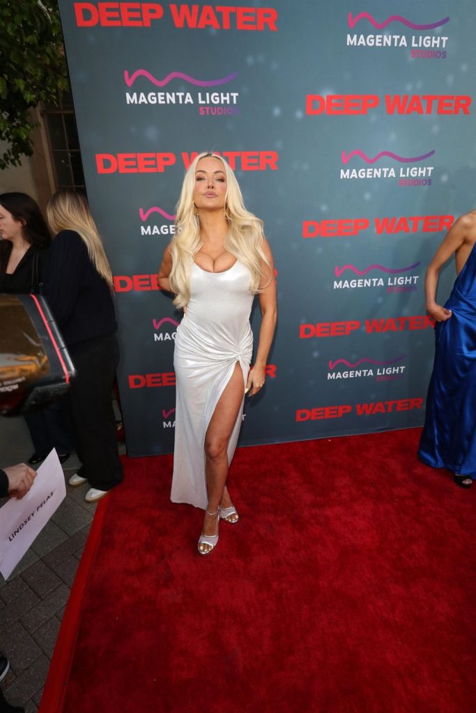 A woman in a white dress with a high slit poses on a red carpet at an event for Deep Water. The backdrop displays logos for Deep Water and Magenta Light Studios in pink and red. Other people stand nearby #2.