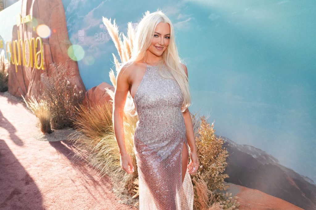 A woman with long platinum blonde hair in a shimmering silver gown stands outdoors on a red carpet, surrounded by desert plants and rocks, with a blue backdrop and sunlight creating lens flares.