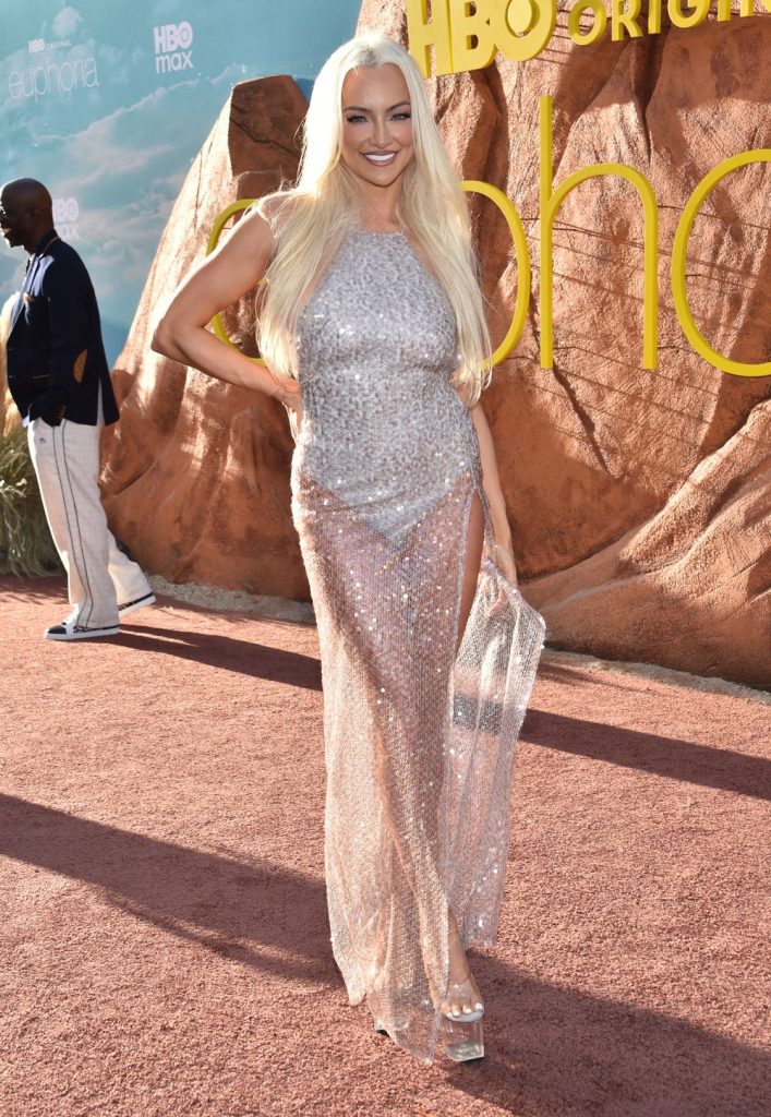 Blonde woman poses on a red carpet in a sparkly, silver dress. Another woman in a dark blue gown stands in the background.