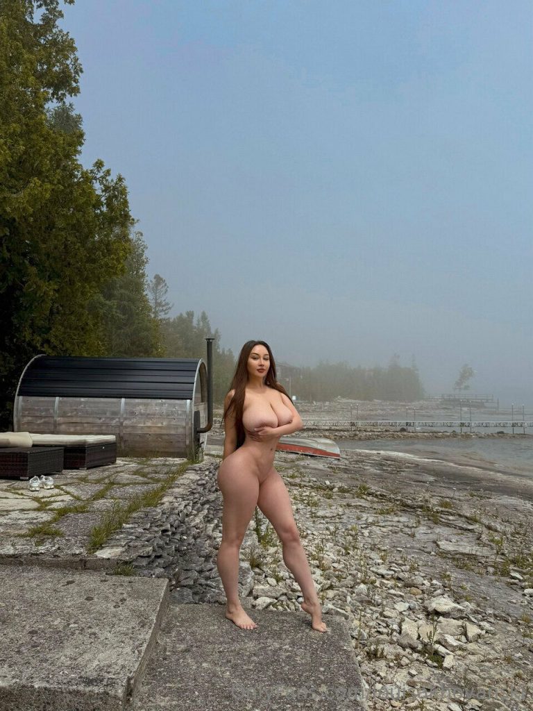 A nude girl standing by the shoreline of a lake.