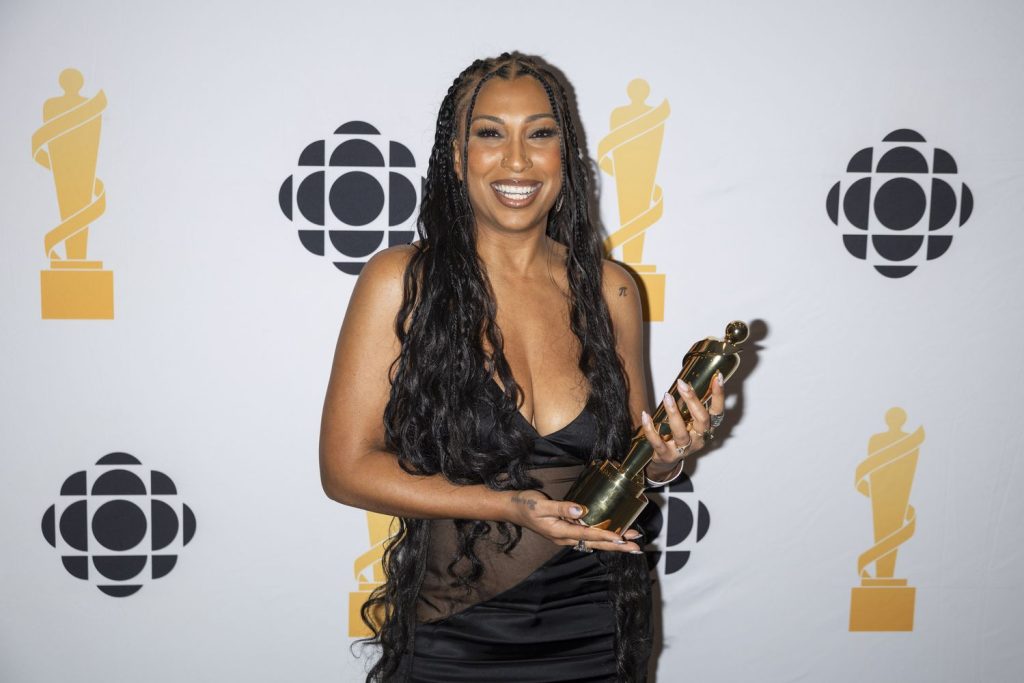 A black woman with long, braided hair smiles while holding a gold trophy. She is wearing a black dress and standing in front of a backdrop with CBC logos and gold award symbols.