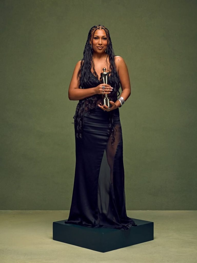 A black woman in a long black dress stands on a platform holding a golden award trophy, with long wavy hair and a confident expression, against a plain green background.