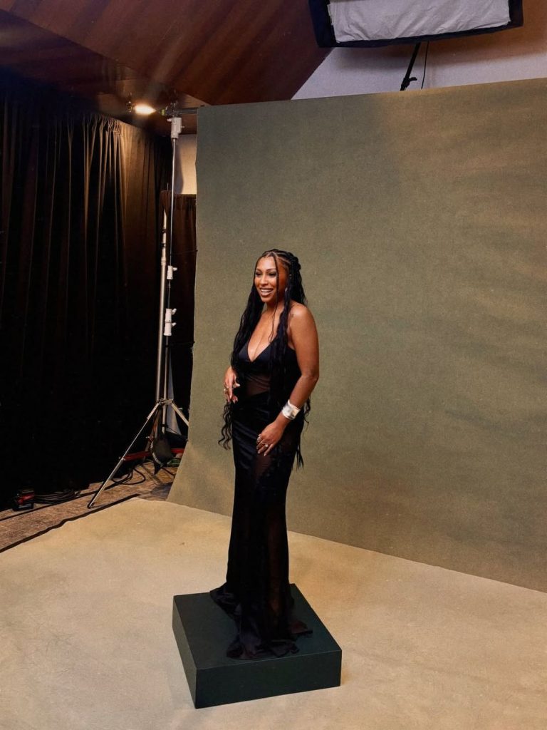 A black woman in a long, black, sleeveless gown stands on a platform in front of a plain green backdrop in a photography studio, smiling at the camera. Studio lights and equipment are visible in the background.