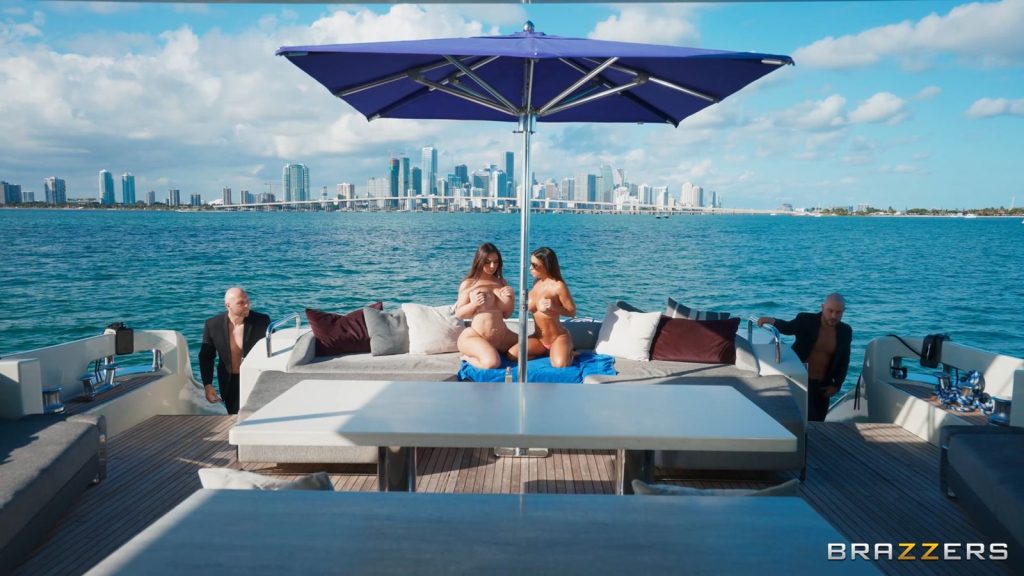 Far away view of two nude women kneeling on a sun bed aboard a boat. Two bald men are seen walking up stairs on both sites of them. Ocean and a city skyline are in the background.