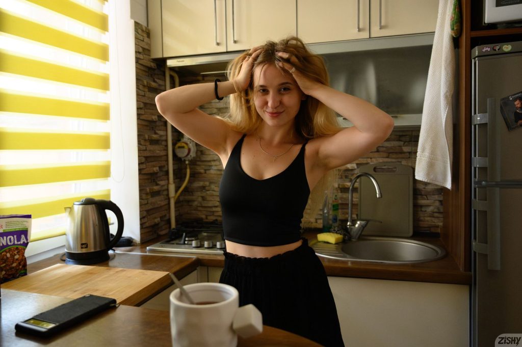 A woman with long blonde hair, wearing a black tank top and skirt, smiles while holding her hands on her head in a modern kitchen with sunlight streaming through yellow blinds. A cup, phone, and cereal box are on the counter.