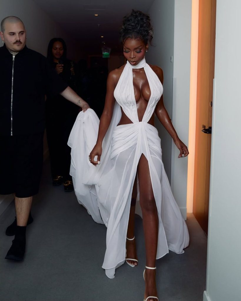 Black woman in a flowing, white, halter-neck gown with cut-out details and a high slit walks down a hallway, holding part of her dress. Two people walk behind her, one dressed in black.