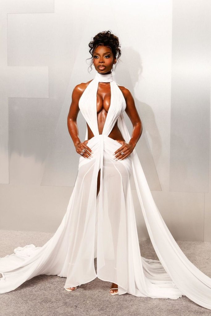 Frontal view of black woman poses confidently in a glamorous, revealing white gown with dramatic draping and cutouts.