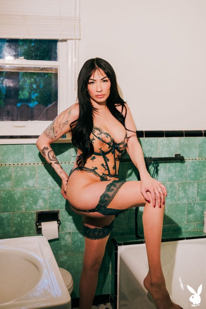 A woman in sheer lace lingerie standing with one leg raised on the edge of a bathtub.