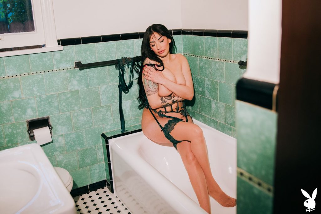 A topless woman wears green lace lingerie and garter belt, sits on the edge of a bathtub in a retro-style bathroom with green tiles.