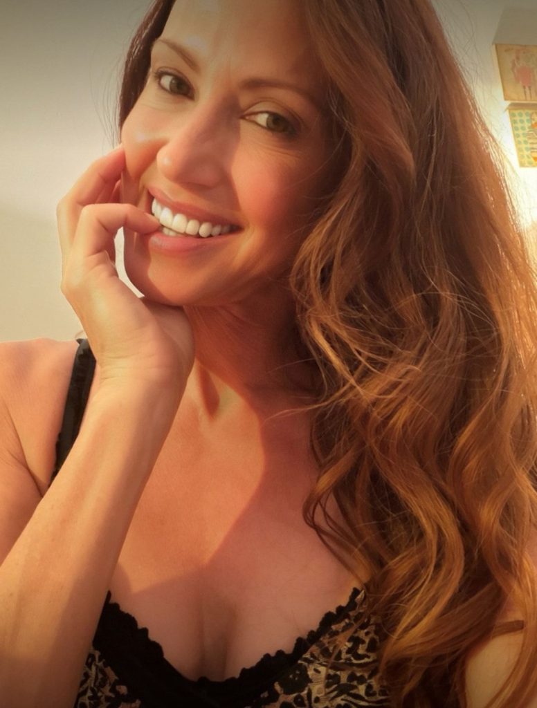 A woman with long, wavy brown hair smiles at the camera, resting her hand on her cheek. She is wearing a sleeveless, patterned top and appears to be in a warmly lit indoor setting.