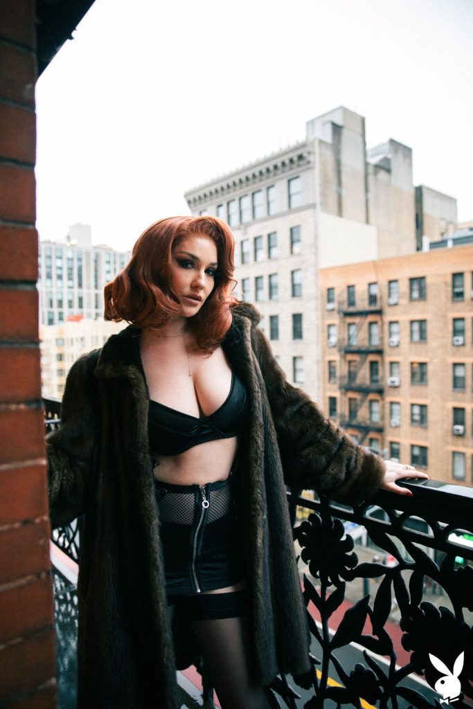 A woman with red hair wearing a dark fur coat, black crop top, and black skirt stands on a balcony overlooking a cityscape with brick buildings.