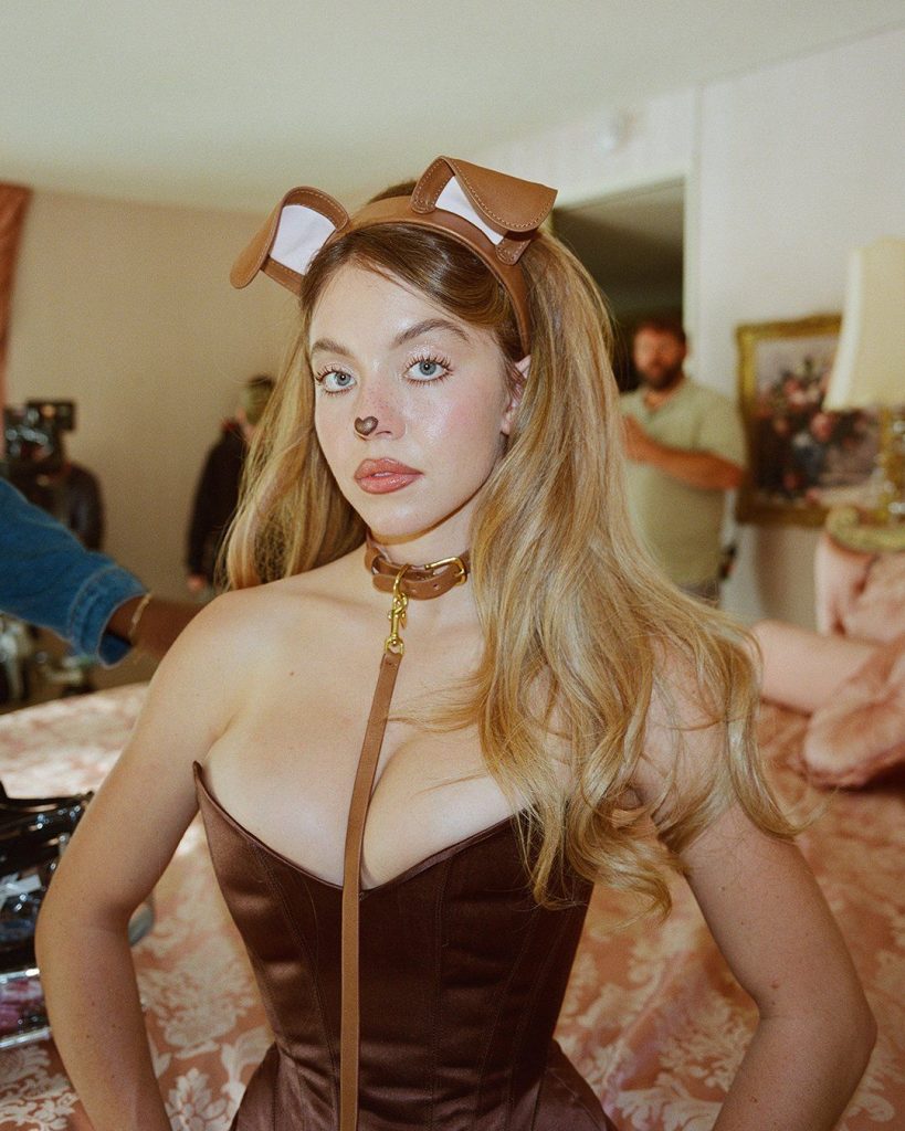 Sydney Sweeney dressed in a brown dog costume, with a headband featuring dog ears, a dog nose painted on her face, and a brown collar with a leash, stands in a room with a pink bedspread and people in the background.