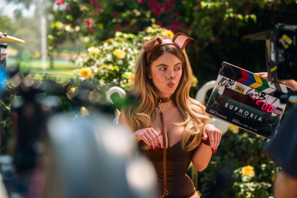 Sydney Sweeney dressed in a brown dog costume poses with her hands bent like paws, making a playful face. Filming equipment and a clapperboard labeled “Euphoria” are visible around her, and yellow flowers are in the background.