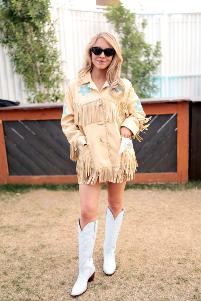 A woman wearing a light tan fringe jacket with blue flower designs, matching fringe mini skirt, white cowboy boots, and sunglasses stands outdoors on grass in front of a wooden fence and greenery.
